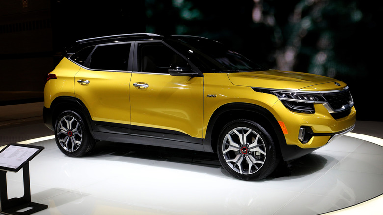 A yellow metallic Kia Seltos for the 2025 model year on display indoors on a podium in a dark room.