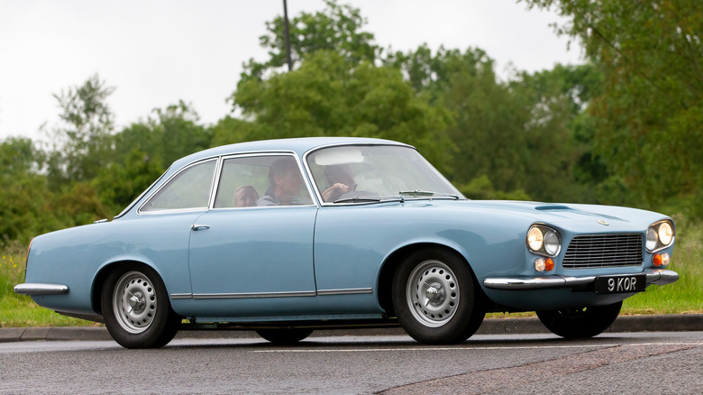 A one of 99 light blue Gordon Keeble grand touring car driving in the rain, trees in the back.